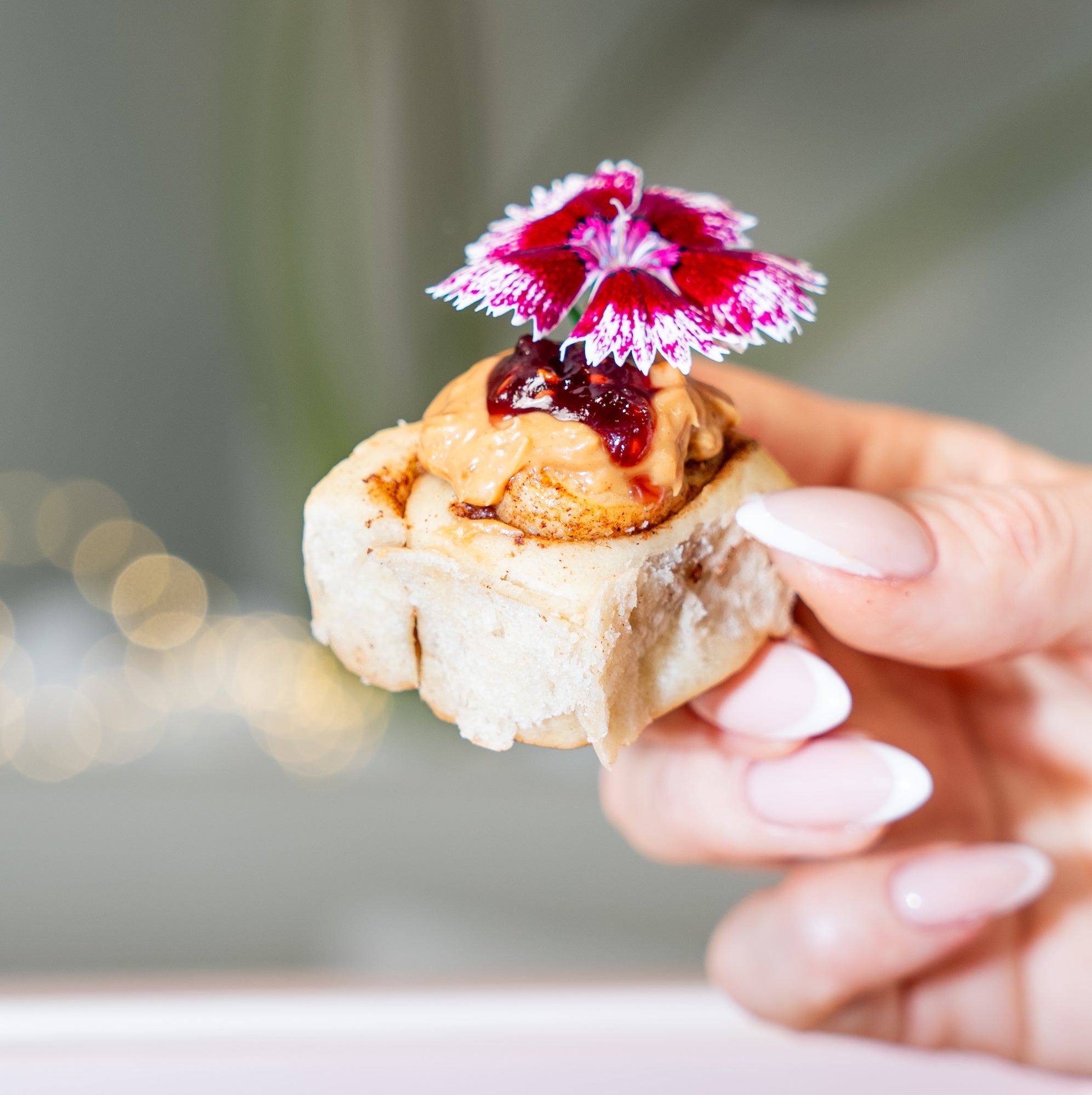 Hand holding a mini cinnamon roll with peanut butter jam and a flower on top against a blurred background