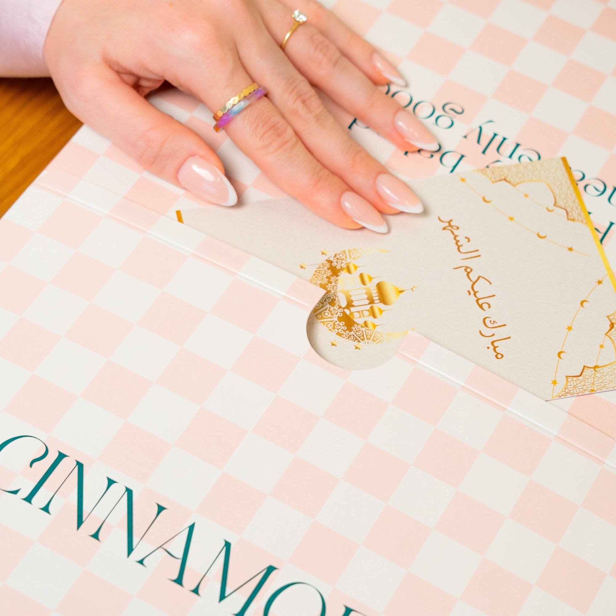 Hand with rings on a pink checkered cinnamon roll box labeled 'CINNAMORE'.